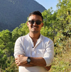 Junger Mann schaut frontal in die Kamera. Er trägt ein weisses Hemb und eine Sonnenbrille. Im Hintergrund sind Bäume zu sehen